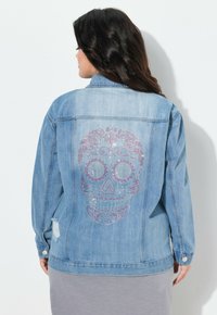 Vaaleansininen farkkutakki, jossa on suuri koristeellinen pääkallo-design vaaleanpunaisista ja hopeisista strasseista selän puolella, sekä kuluneet yksityiskohdat.