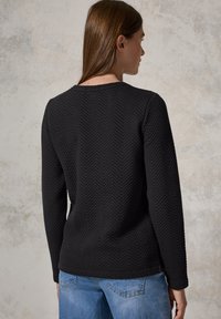 Svart långärmad topp med en strukturerad herringbone-mönster, rundad halsringning och rak passform, i kombination med ljusblå jeans.