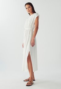 Bílá, volná maxi šaty s krátkými, strukturovanými rukávy, elastickým pasem a bočními rozparky. Nosené s černými sandály na tenké pásky na jednobarevném pozadí.