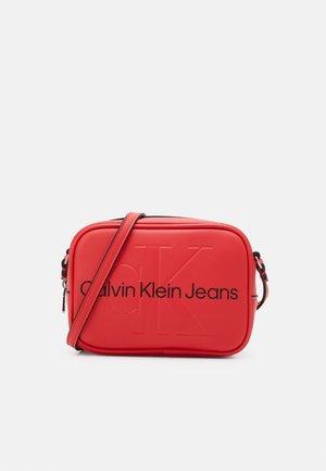 Czerwona prostokątna torba na ramię z wytłoczonym logo "CK" i czarnym napisem "Calvin Klein Jeans" na przodzie, regulowany pasek na ramię oraz zapięcie na zamek błyskawiczny.
