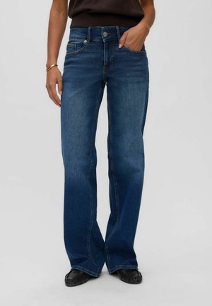 Person iført mørkeblå jeans med vide ben og forlommer, sorte loafers, en sort top og et guldarmbånd på venstre håndled.