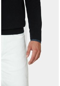 Marineblaues Strickpullover mit gerippten Bündchen, kombiniert mit weißen Hosen. Der Pullover hat ein kontrastierendes blaues Innenbündchen.