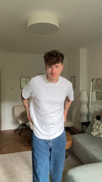 T-shirt branca de manga curta combinada com jeans denim azuis. O cenário apresenta um ambiente claro com móveis e decoração moderna.