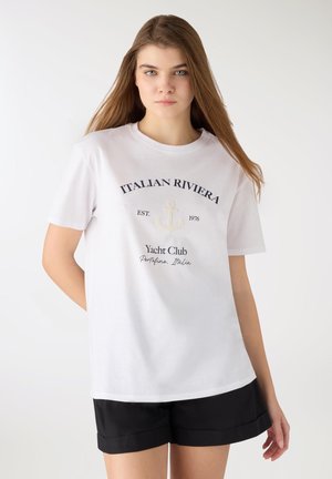 Giovane donna con capelli lunghi che indossa una T-shirt bianca con la scritta "Italian Riviera Yacht Club" e pantaloncini neri, in piedi davanti a uno sfondo neutro.