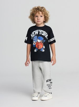 T-shirt nera con grafica "NEW YORK", pantaloni della tuta grigi con stampa testuale, sneaker bianche, capelli ricci. Outfit sportivo casual.