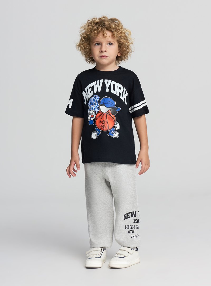 Czarna koszulka z nadrukiem "NEW YORK", szare spodnie dresowe z nadrukiem tekstowym, białe trampki, kręcone włosy. Codzienny sportowy strój.