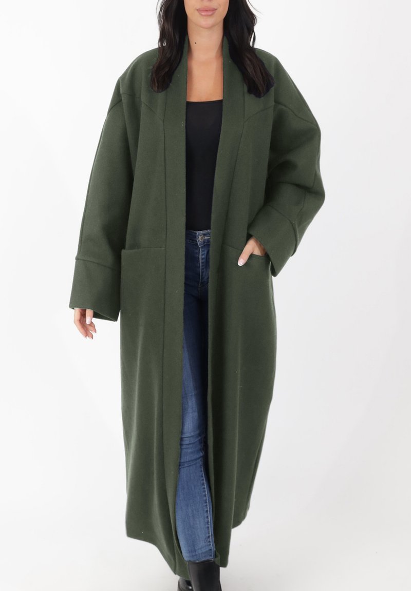 Lange jas met open voorkant in donker groene stof; heeft voorzakken, brede mouwen en een ontspannen pasvorm, gedragen over een zwarte top en spijkerbroek.