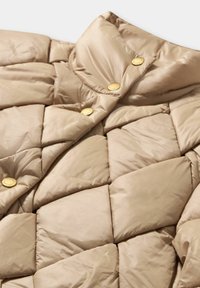 Beżowa kurtka puchowa z pikowanym wzorem, wyposażona w wysoki kołnierz i okrągłe złote guziki. Miękki, błyszczący materiał o gładkiej fakturze.