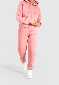 Sudadera con cremallera en rosa claro, con cordones y logo blanco, combinada con pantalones de chándal de cintura elástica a juego y zapatillas blancas.