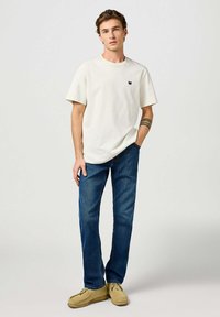 Camiseta de algodón color crema claro con mangas cortas y un pequeño logo negro, combinada con jeans rectos de color azul oscuro y zapatos mocasín beige.