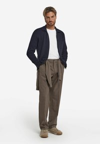 Marinblå stickad cardigan över en vit T-shirt, kombinerad med lösa bruna rutiga byxor och beige sneakers, med en avslappnad knytning i midjan.