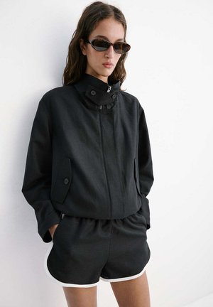 Femme portant une veste noire à col haut avec des poches boutonnées et un short noir avec un bord blanc, debout contre un mur blanc, mains dans les poches.
