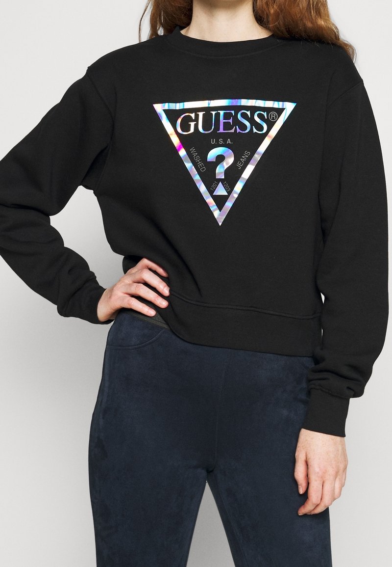 Personne portant un sweat-shirt noir Guess avec un logo holographique et un pantalon bleu foncé, main posée sur la hanche sur fond clair.