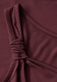Burgundy ärmlös topp med samlad remdetalj, slät textur och rund halsringning. Materialet verkar mjukt och elastiskt.