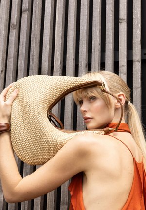 Frau mit blonden Haaren und orangefarbenem Oberteil hält große, gewebte, halbmondförmige Tasche vor einem Hintergrund aus Holzlatten.