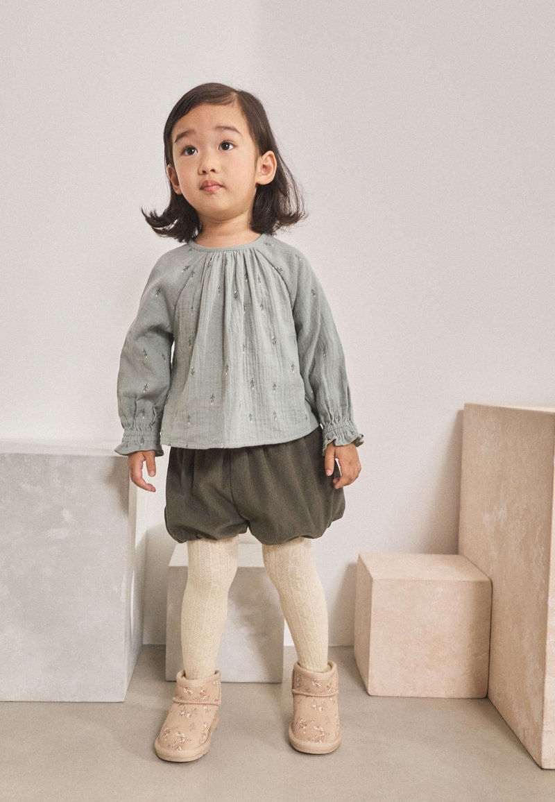 Chemise à manches longues vert clair avec broderie florale, associée à un short vert foncé, des collants crème texturés et des bottines beige avec des accents floraux.