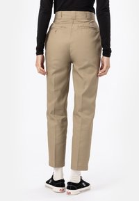 Pantalon beige à taille haute, avec poches latérales et poches arrière. Tissu lisse avec une coupe ajustée et ourlets aux chevilles. Chaussures noires portées avec des chaussettes blanches.