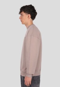 Svetlo bež majica z okroglim izrezom in rebrastimi manšetami, z nežnim logotipom na prsih in sproščenim krojem.