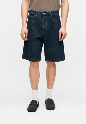 Mann trägt dunkle Denim-Kniehosen, weiße Socken, schwarze Slip-On-Schuhe und ein braunes T-Shirt.