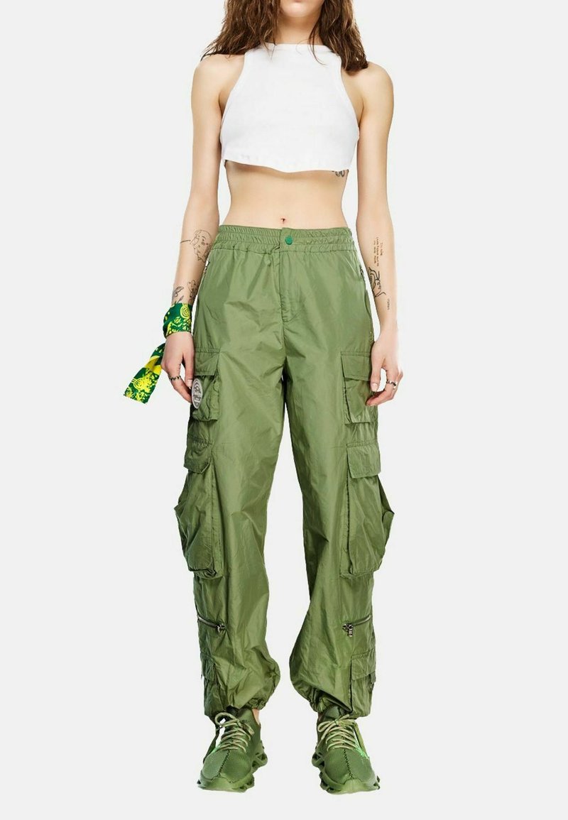 Γυναίκα που φοράει λευκό crop top, πράσινο παντελόνι cargo με πολλές τσέπες, πράσινα αθλητικά παπούτσια και πράσινο μαντήλι με σχέδια στον καρπό.