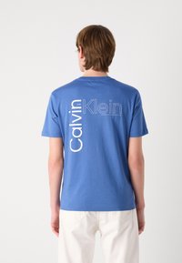 Blue cotton t-shirt featuring the text "Calvin Klein" in white vertical lettering on the back, crew neck, short sleeves, and relaxed fit.