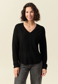 Donna con capelli ricci che indossa un maglione nero a scollo a V, jeans grigi e collane d'oro a strati, sorridente su uno sfondo beige.