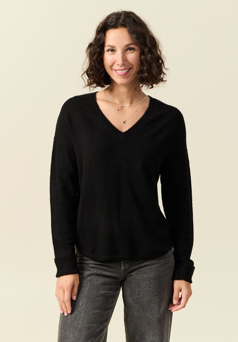 Donna con capelli ricci che indossa un maglione nero a scollo a V, jeans grigi e collane d'oro a strati, sorridente su uno sfondo beige.