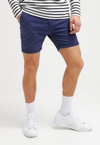 Námořnické bavlněné šortky s uvolněným střihem, ohrnutými nohavicemi a bočními kapsami, v kombinaci s bílými teniskami a bílými ponožkami do kotníků.