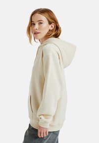 Beige hoodie met een grote capuchon, kangoeroezak en geribde manchetten. De stof lijkt zacht en textuur te hebben, met een ontspannen pasvorm.