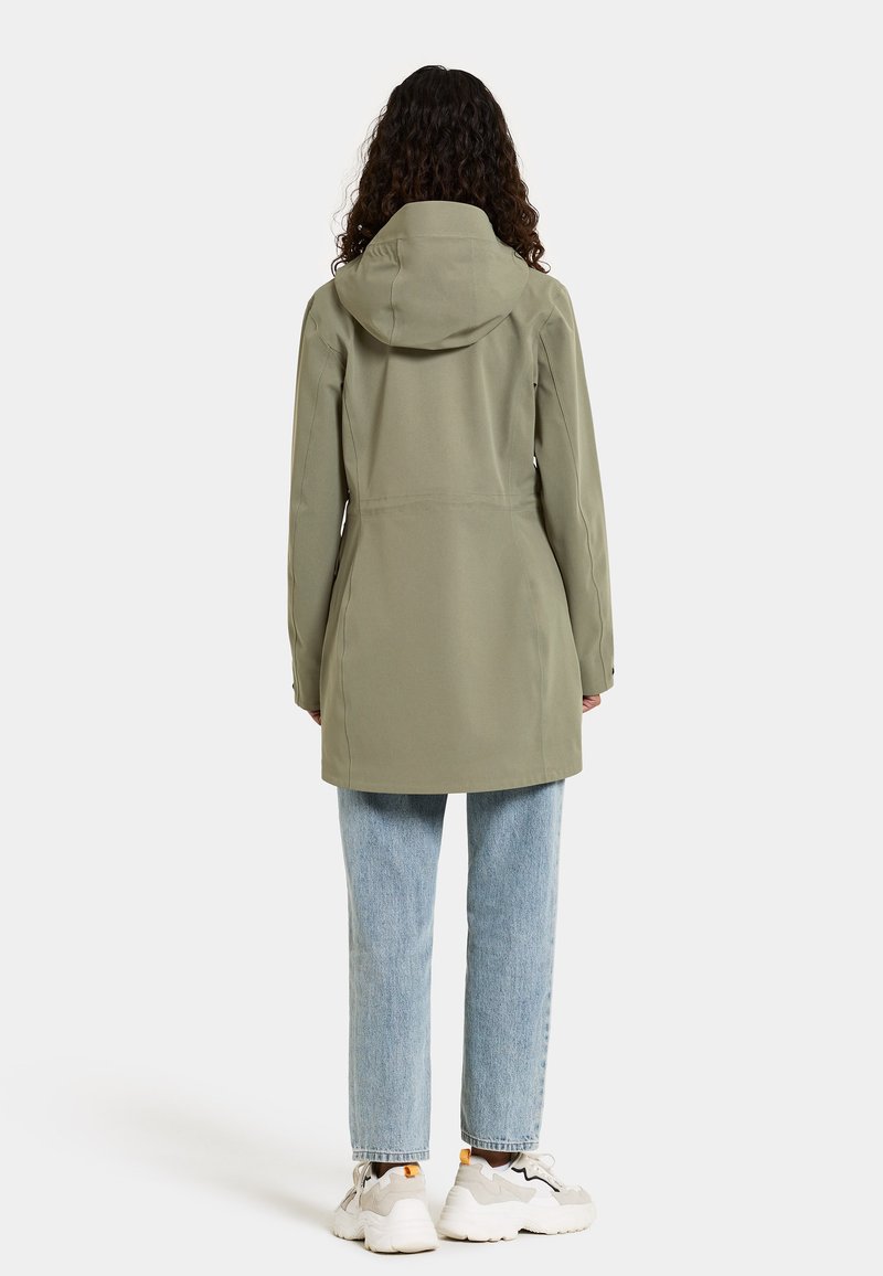 Olivgrüne wasserdichte Jacke mit Kapuze, mittellanges Design und glatter Textur, kombiniert mit hellblauen Jeans und klobigen Sneakern.