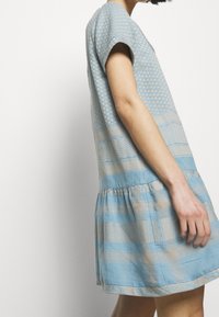 Světle modré šaty s krátkými rukávy a sukni s volány, které mají tkané vzory v ještě světlejší modré a béžové barvě. Vyrobeno z měkké látky.