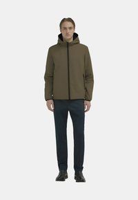 Giacca con cappuccio color kaki con zip frontale e tasche laterali, abbinata a pantaloni blu navy e scarpe nere, indossata da una figura in piedi.