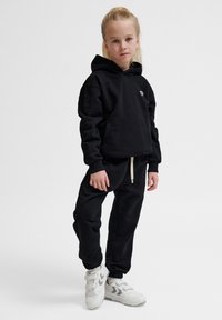 Schwarzes Hoodie- und Jogginghosen-Set in lockerer Passform. Das Hoodie hat eine Kängurutasche und ein Logo; die Hose hat elastische Bündchen und einen Kordelzug. Weiße Sneaker.