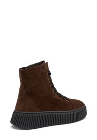 Botas de ante marrón hasta el tobillo con cordones negros, ojales dorados y una suela de goma negra texturizada. Presenta una punta redondeada y un diseño de caña alta.