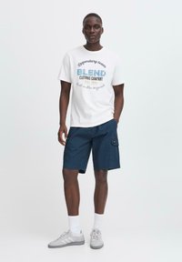 Baltas t-kreklis ar "BLEND" logotipu zilā un pelēkā krāsā, kombinēts ar tumši ziliem šortiem. Modelis valkā pelēkas sporta kurpes un baltas zeķes.