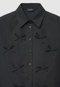 Schwarzes Hemd zum Knöpfen mit spitzem Kragen und dekorativen Stoffschleifen, die auf der Vorderseite angeordnet sind. Das Etikett trägt die Aufschrift "CALLIOPE S."