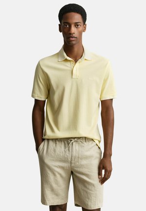 Man die een lichtgeel poloshirt met korte mouwen en beige shorts met trekkoord draagt, staat met handen in de zakken tegen een witte achtergrond.
