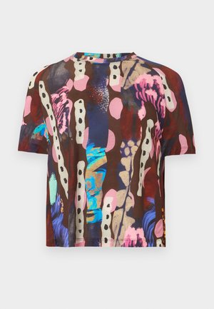 Moteris stovi, rankas kišenėse, dėvi trumpomis rankovėmis marškinius su margu abstrakčiu raštu, juodus kelnes ir rudus batus.