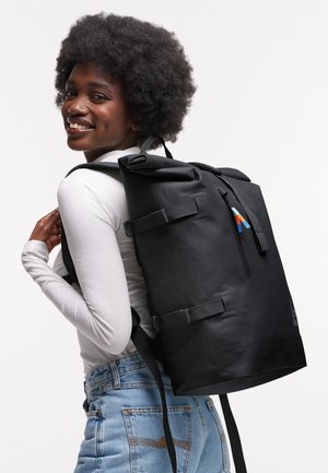 Hymyilevä nainen kiharat hiukset, valkoinen pitkähihainen paita ja farkut, kantaa suurta mustaa reppua selässään.