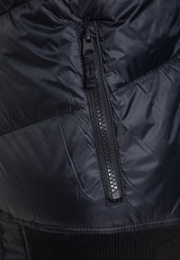 Detalje af sort dunjakke med lynlåslomme, skinnende tekstur og ribstrikket kant. Quiltet mønster synligt på stofoverfladen.