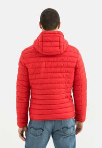 Rote gesteppte Pufferjacke mit Kapuze, mit horizontalen Nähten, Reißverschluss vorne und elastischen Bündchen. Tragen mit blauen Jeans.