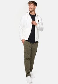 Weißer Zip-Hoodie mit Kordelzügen, über einem schwarzen Shirt getragen, kombiniert mit olivfarbenen Cargo-Hosen und weißen Sneakers. Lässiges Outfit-Design.