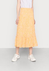 Saia midi amarela com padrão floral, design em camadas e bainha com ondas, confeccionada em tecido leve. Usada com ténis brancos.