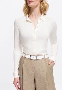 Femme portant une chemise blanche à manches longues rentrée dans un pantalon en lin beige taille haute avec une ceinture blanche et une main dans la poche.