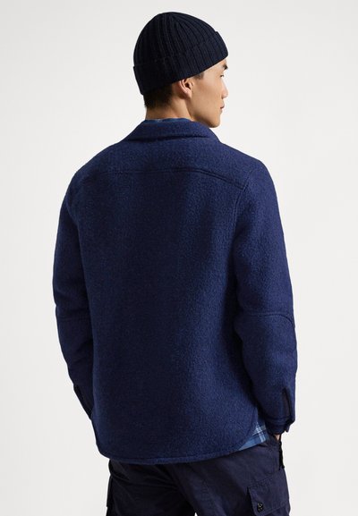 Marineblaue Fleecejacke mit strukturierter Oberfläche, ausgestattet mit spitzem Kragen und Seitentaschen. Kombiniert mit einer passenden gerippten Strickmütze.