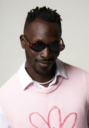 Roze gebreide trui met een rood bloemenpatroon, gedragen over een witte blouse met kraag. Bruine ovale zonnebril en een parelhoofdsieraad maken de look af.