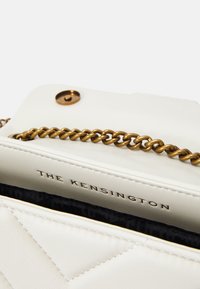 Vitt läderhandväska med ett kviltat mönster, guldfärgad kedjestrap, och präglad "THE KENSINGTON"-logotyp på det inre fodret.