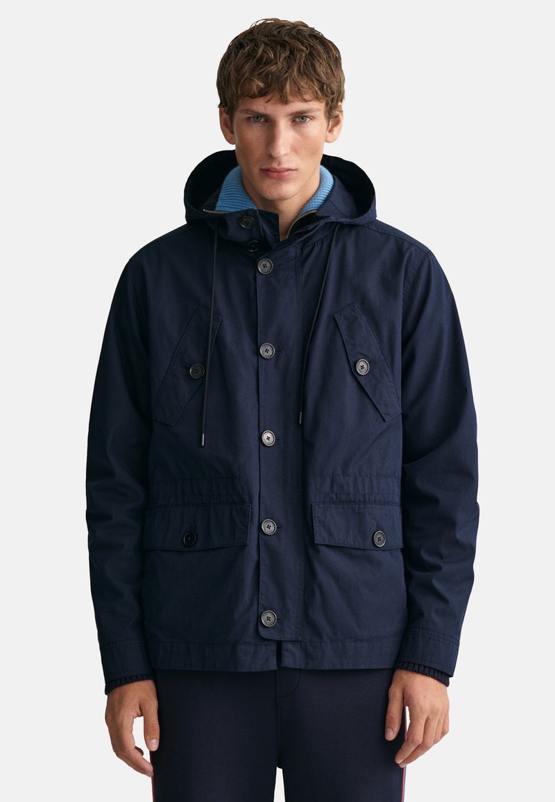 Granatowa kurtka z kapturem, przednimi guzikami, podwójnymi kieszeniami na piersi i elastycznymi mankietami. Wykonana z trwałej, teksturowanej tkaniny.