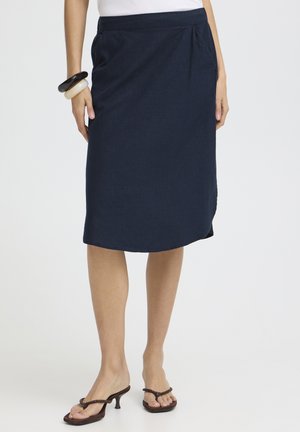 Femme portant une jupe bleu marine longueur genou avec poches latérales, un haut blanc, des bracelets épais et des sandales à talons marron.