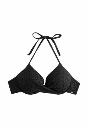 Femeie în costum de baie negru, stând pe o plajă nisipoasă cu apă turcoaz și dealuri verzi în fundal, sub un cer parțial înnorat.